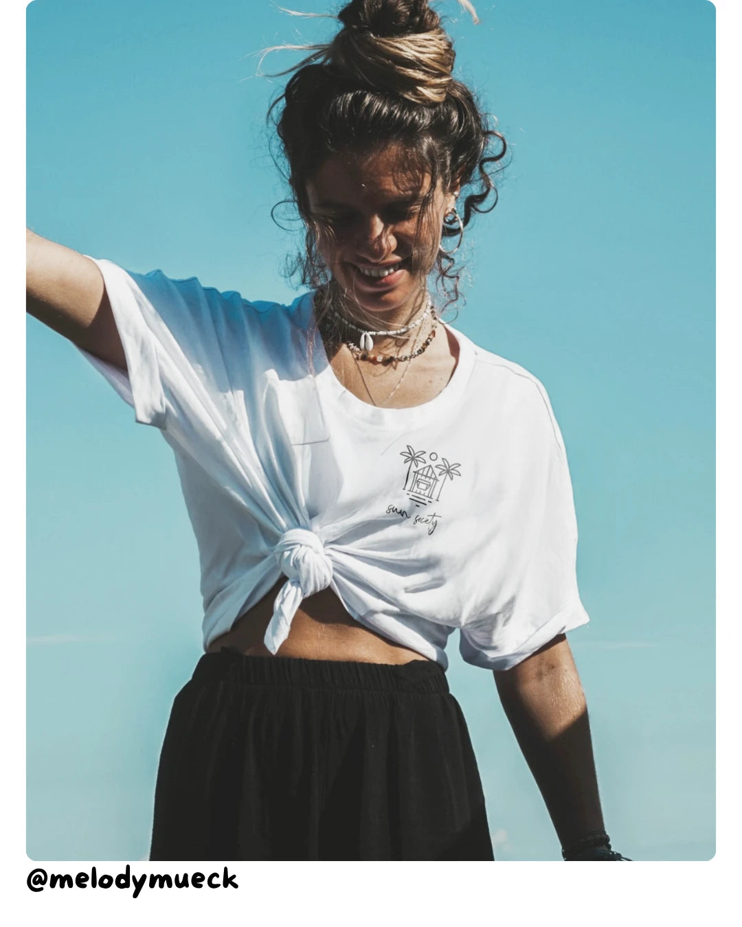 Model von The Sunkissed Rebels trägt ein weißes Sommer-T-Shirt im Outdoor-Look vor strahlend blauem Himmel.