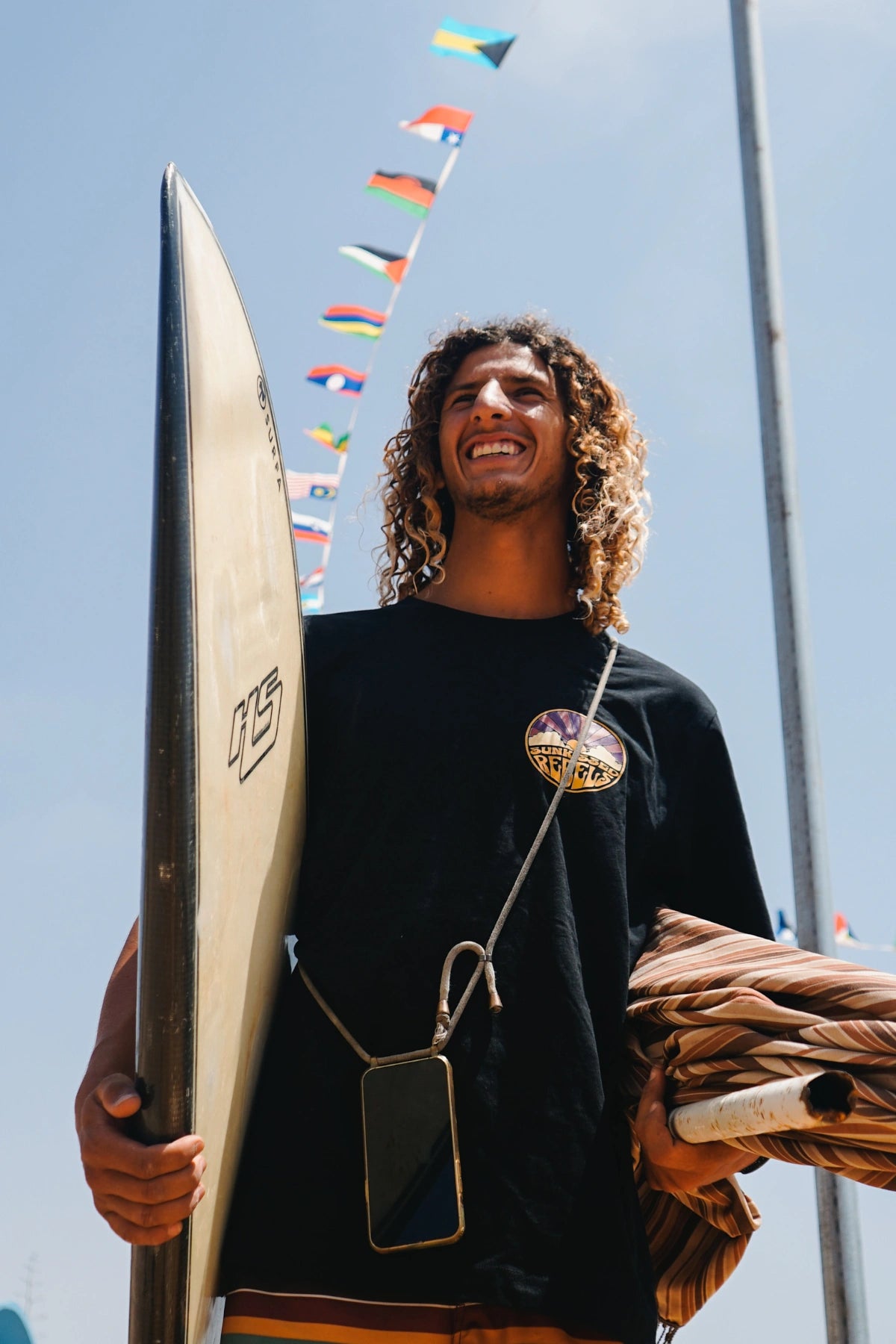 Surfer blacht in Kamera und hälkt Surfbrett unterm Arm. Dabeiu trägt er ein schwarzesy Oversized T-Shirt von The Sunkissed Rebels