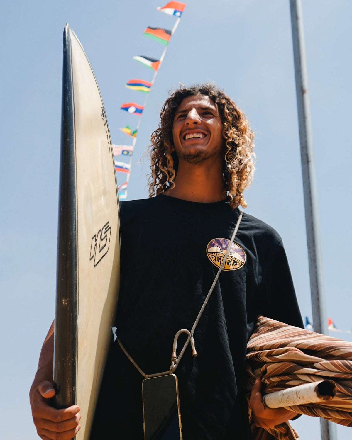 Surfer blacht in Kamera und hälkt Surfbrett unterm Arm. Dabeiu trägt er ein schwarzesy Oversized T-Shirt von The Sunkissed Rebels