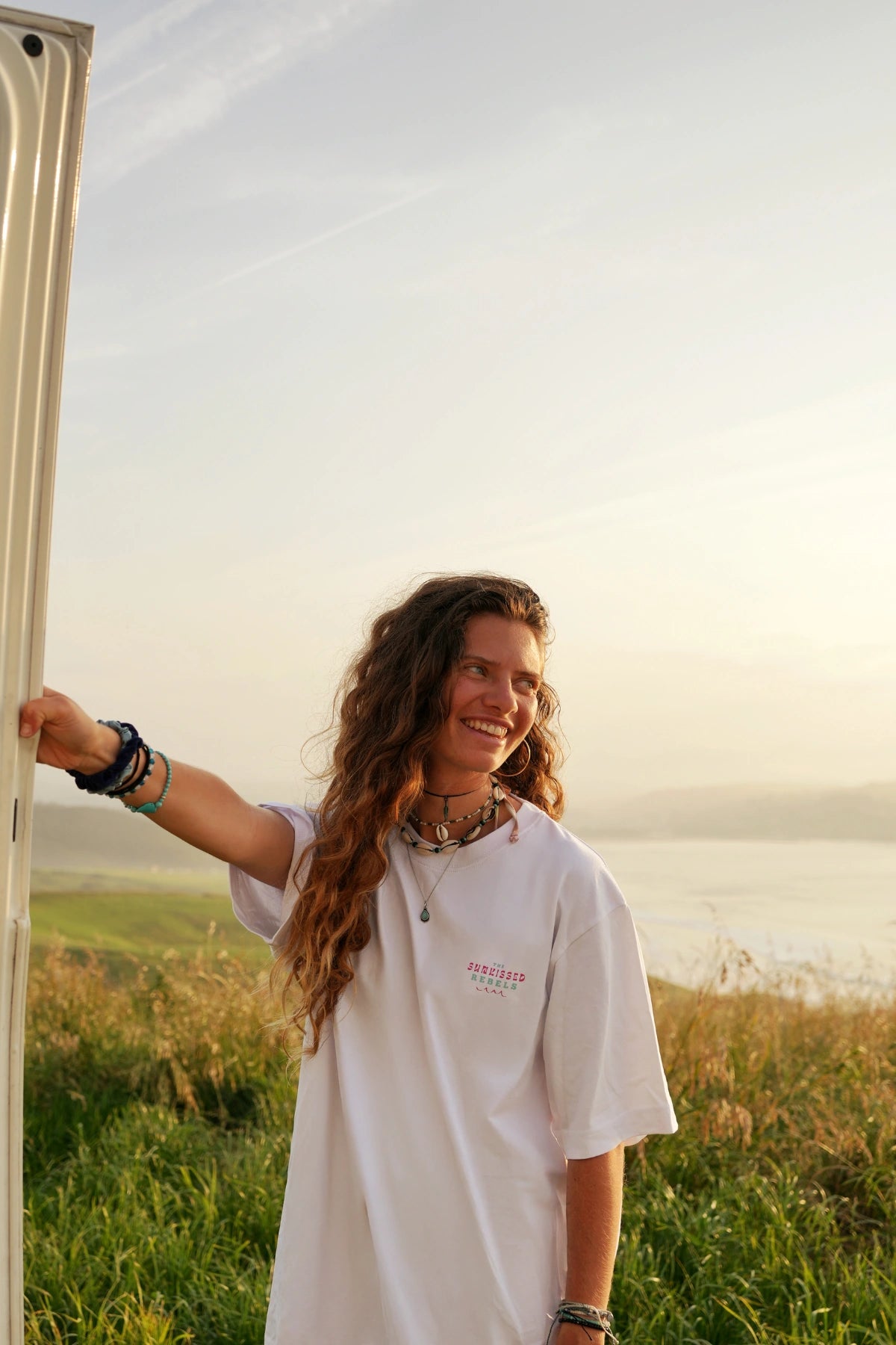 Frau in weißem Oversized Shirt mit Schmuck lächelt entspannt in die Sonne – Freiheitsmoment pur