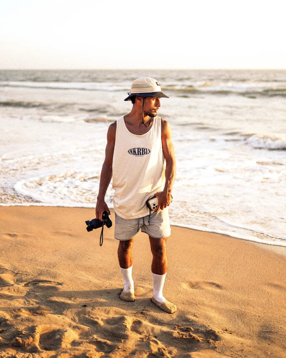 Mann am Strand mit Kamera im SNKRBLS Tank Top Natural Raw – lässiger Sunset-Look mit Bucket Hat