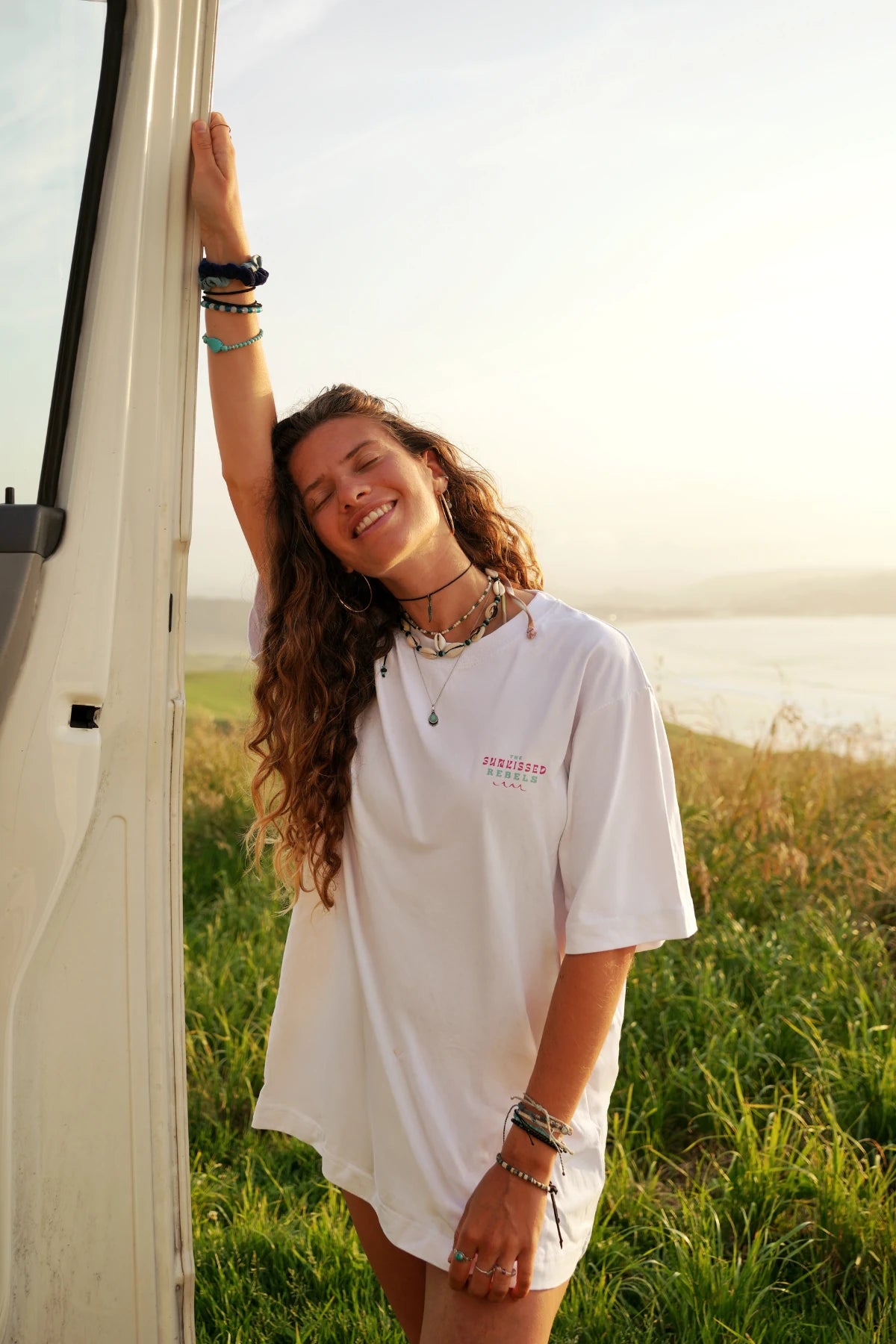 Frau im weißen Vacation Vibes Oversized T-Shirt lächelt entspannt im Sonnenuntergang am Camper