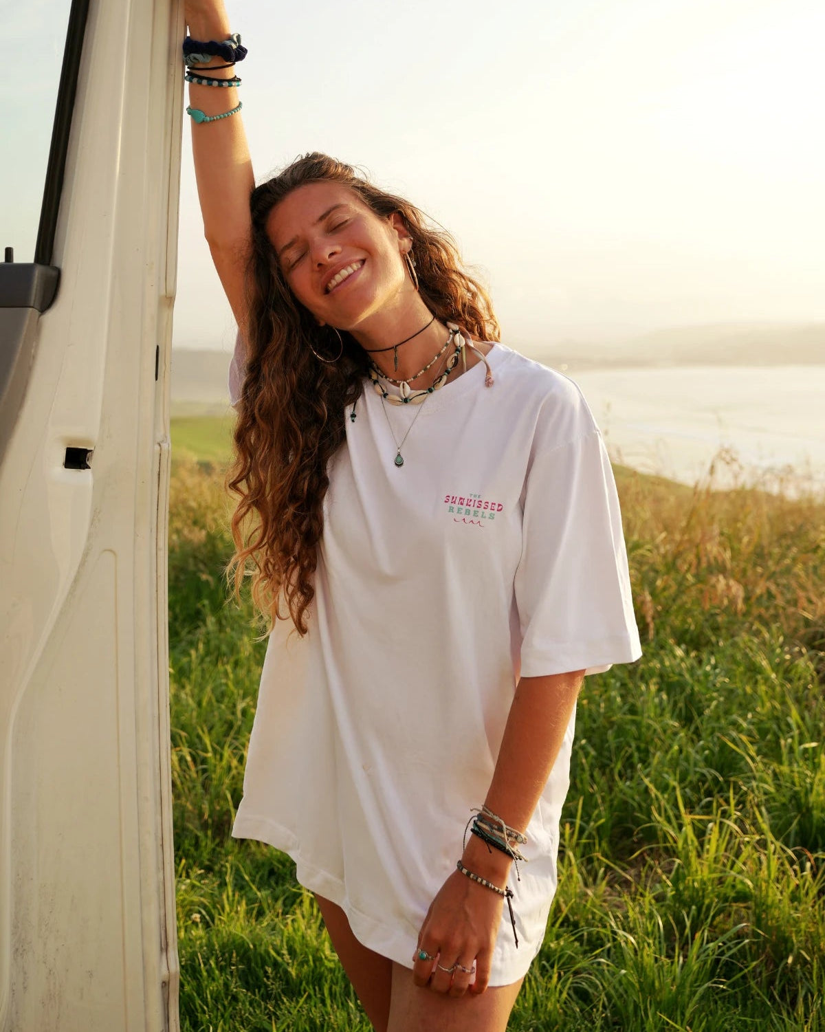 Frau im weißen Vacation Vibes Oversized T-Shirt lächelt entspannt im Sonnenuntergang am Camper