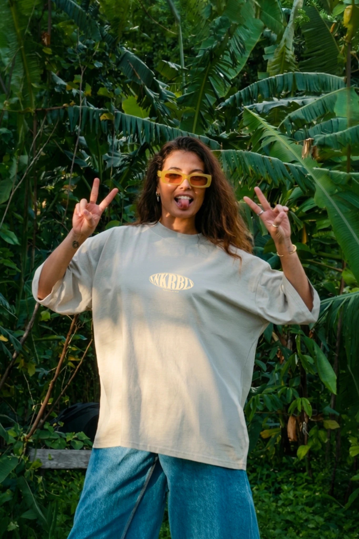„Frau mit Sonnenbrille und oversize dT-shirt in stone zeigt Peace-Zeichen im tropischen Dschungel – lässiger Sunset Rebel Lifestyle in der Natur“