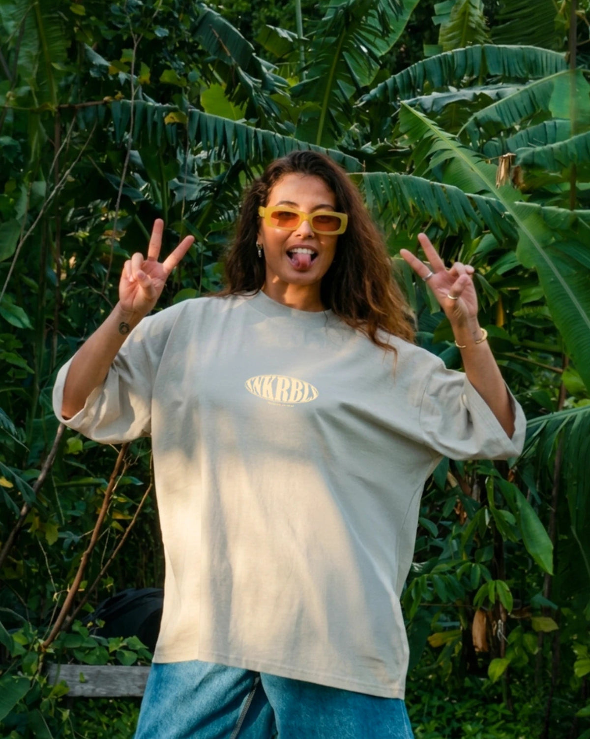 „Frau mit Sonnenbrille und oversize dT-shirt in stone zeigt Peace-Zeichen im tropischen Dschungel – lässiger Sunset Rebel Lifestyle in der Natur“
