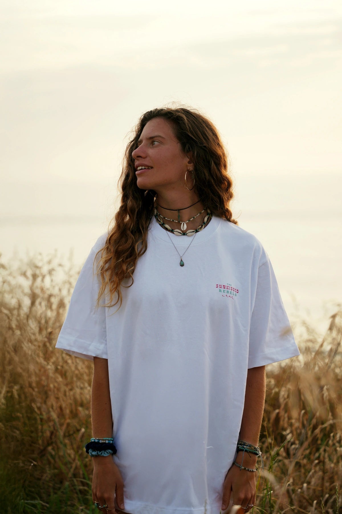 Naturverbundene Frau mit Schmuck und weißem The Sunkissed Rebels T-Shirt vor Meereskulisse im Abendlicht.