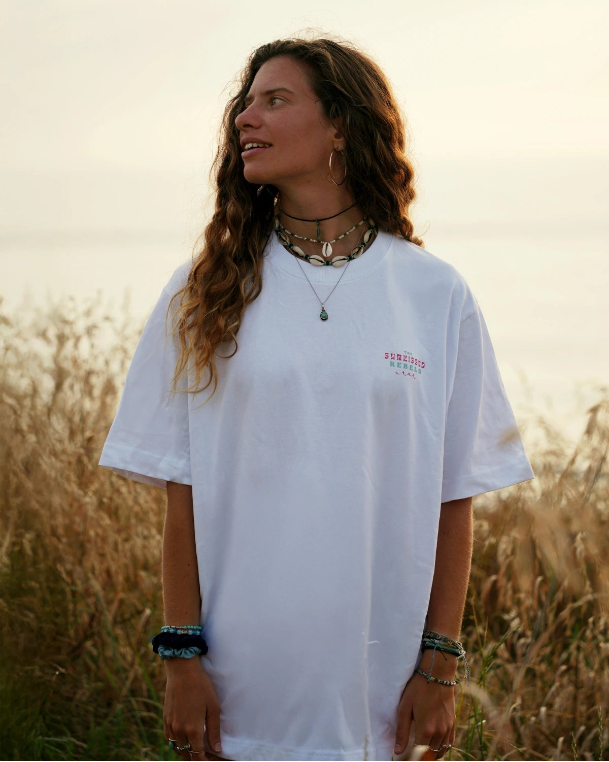 Naturverbundene Frau mit Schmuck und weißem The Sunkissed Rebels T-Shirt vor Meereskulisse im Abendlicht.