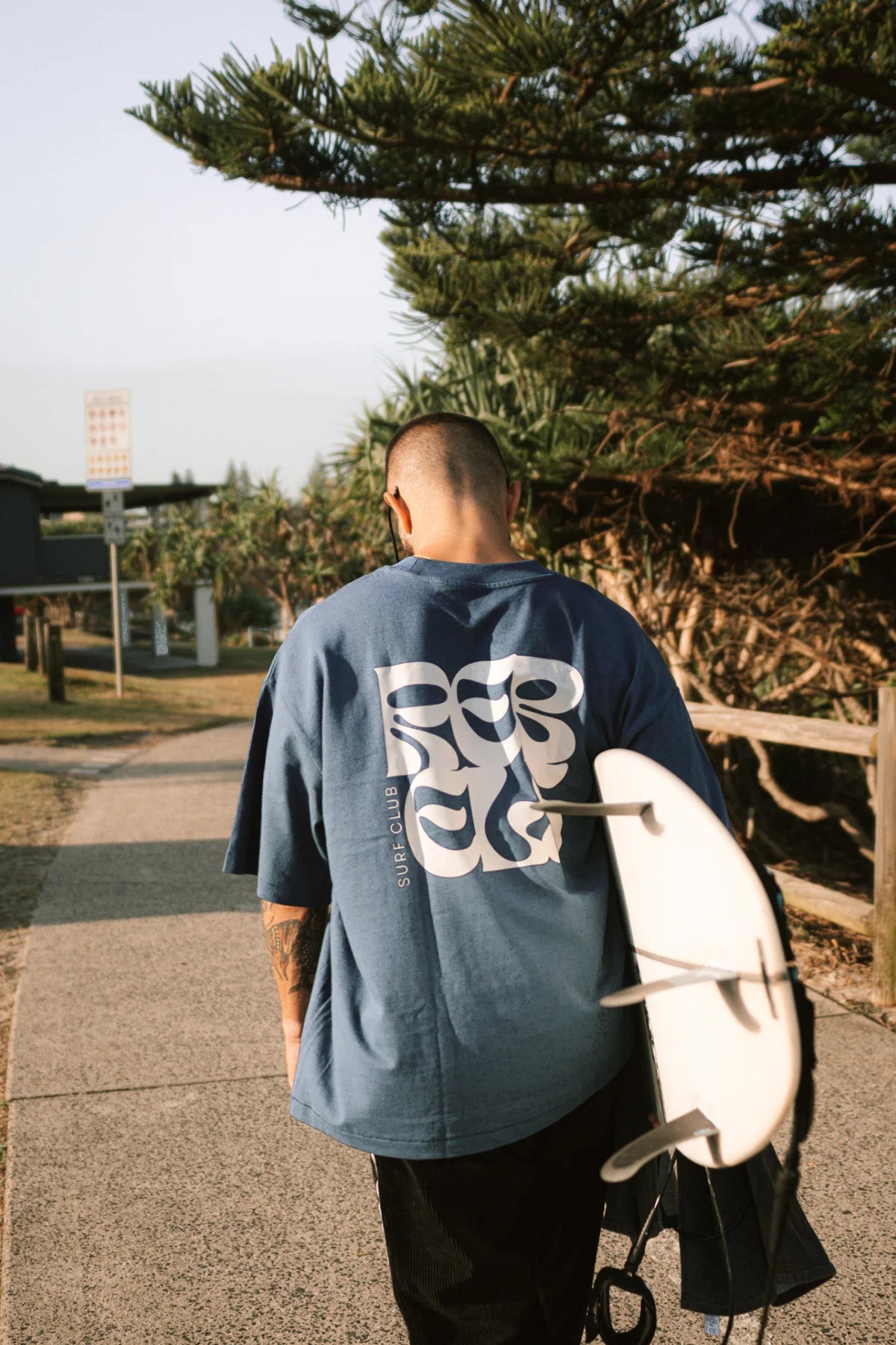 Rückansicht eines Mannes mit Surfbrett im Surf Club Heavy T-Shirt Deep Blue – von The Sunkissed Rebels entspannter Surf Lifestyle am Meer