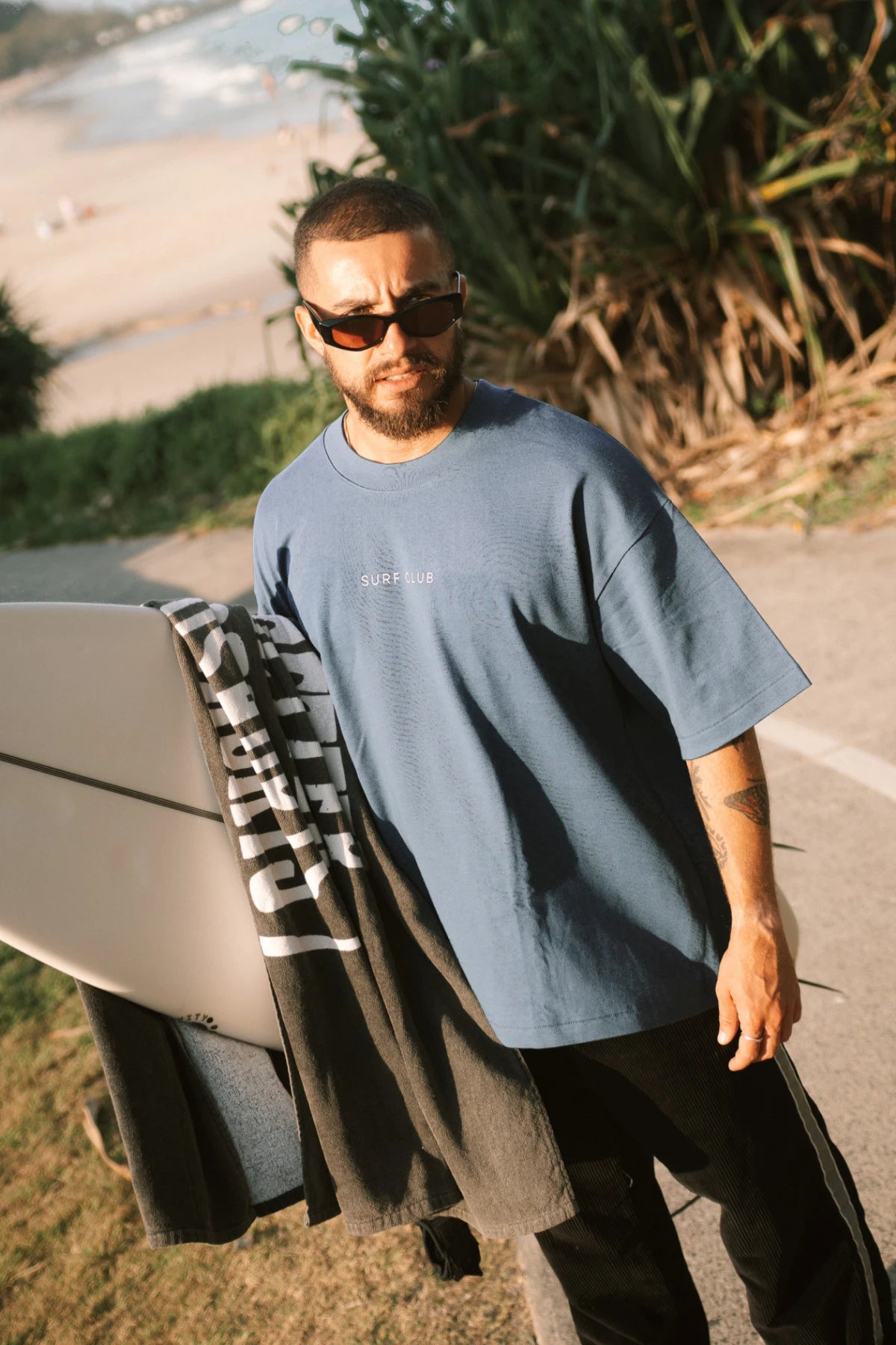 Surfer trägt das Surf Club Heavy T-Shirt – Deep Blue von The Sunkissed Rebels bei Sonnenuntergang am Strand, lässiger Oversized Look für Herrenmode Sommer.