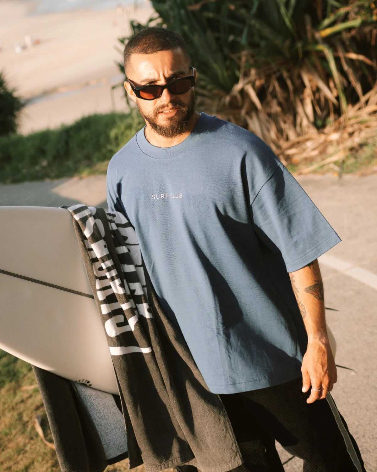 Surfer trägt das Surf Club Heavy T-Shirt – Deep Blue von The Sunkissed Rebels bei Sonnenuntergang am Strand, lässiger Oversized Look für Herrenmode Sommer.