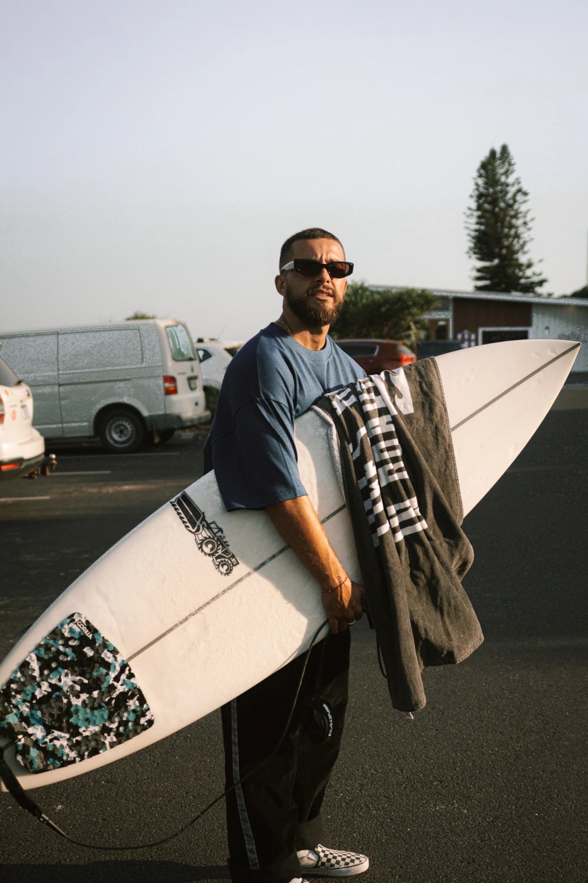 Mann mit Surfbrett und Surf Club Heavy T-Shirt in Deep Blue von The Sunkissed Rebels, sportlicher Surfer Look für Strand und Freizeit.