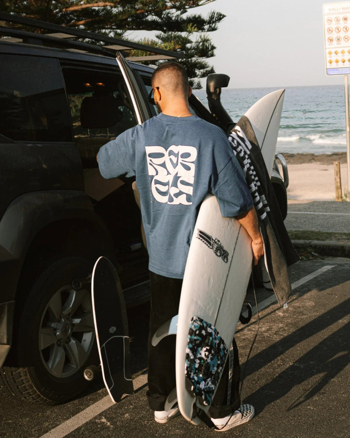 Surf Club Heavy T-Shirt in Deep Blue von The Sunkissed Rebels – Surfer lädt Board in den Van, authentischer Beach Lifestyle Moment.