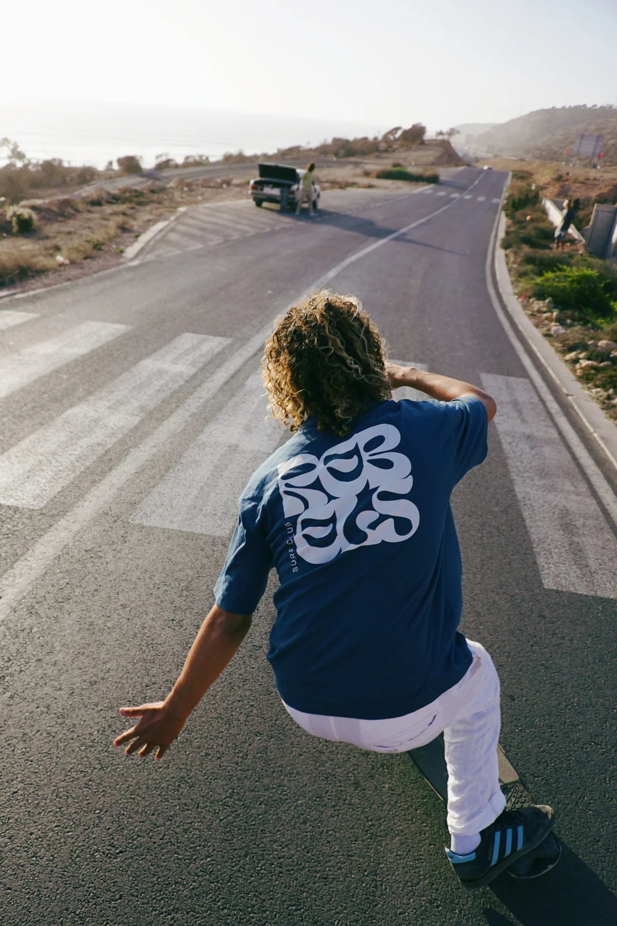 Model von The Sunkissed Rebel fährt dynamisch auf einem Skateboard eine kurvige Straße entlang und trägt das Surf Club T-Shirt in Deep Blue.