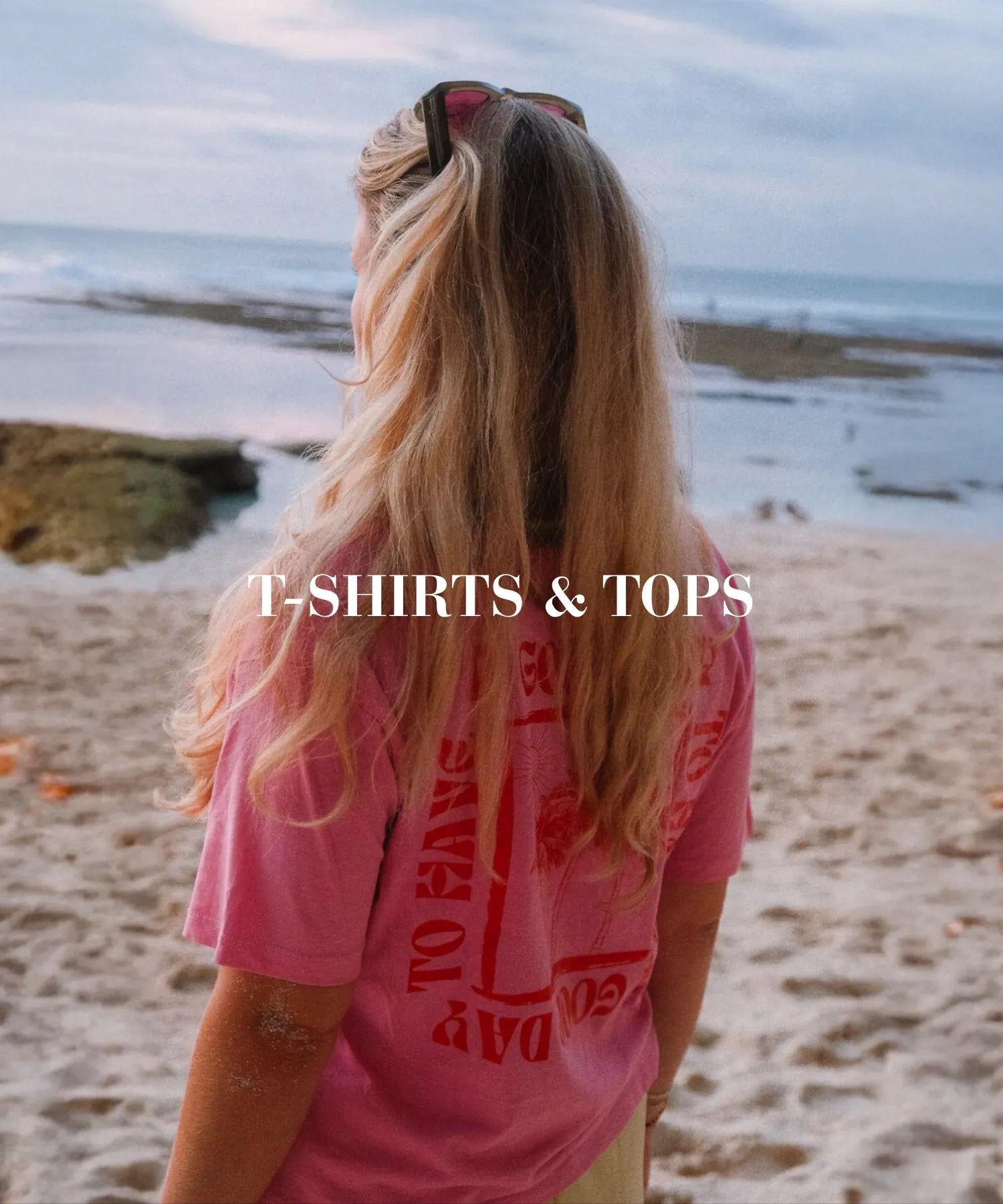 Person mit langem blondem Haar am Strand, trägt pinkes T-Shirt mit Rückenprint, Meereskulisse bei Sonnenuntergang – Kategorie T-Shirts & Tops