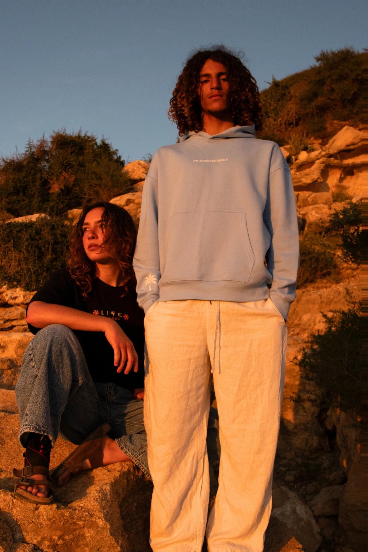 Zwei Models in nachhaltiger Casual Wear bei Sonnenuntergang, mit Fokus auf den hellblauen The Horizon Hoodie Blue Soul.