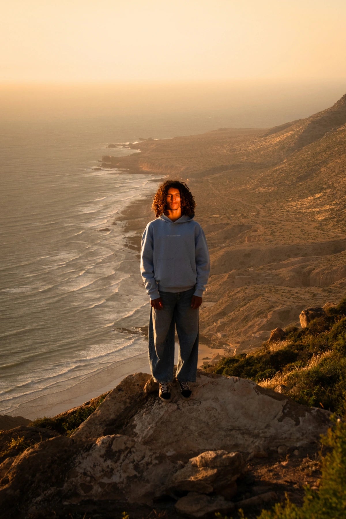 Model trägt den hellblauen The Horizon Hoodie Blue Soul auf einer Klippe mit Blick auf das Meer während eines goldenen Sonnenuntergangs.