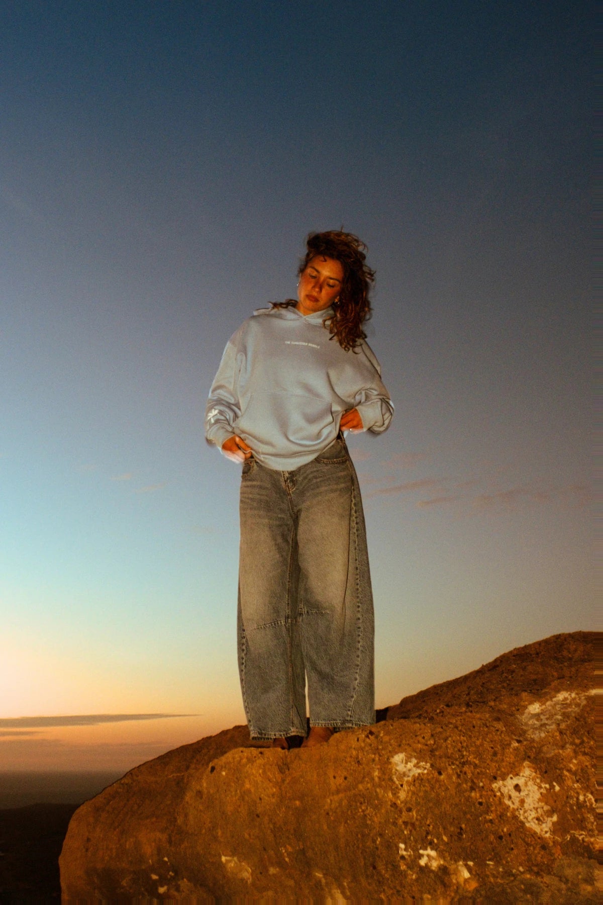 Weibliches Model trägt den hellblauen The Horizon Hoodie Blue Soul auf einer Felsformation im warmen Abendlicht.