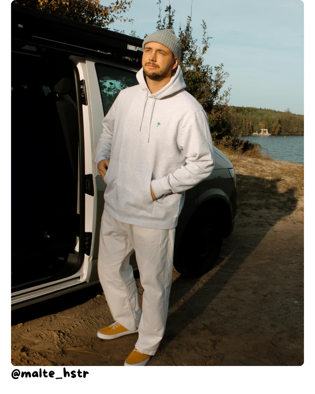 Model von The Sunkissed Rebel trägt einen grauen Hoodie vor einem Campervan am See.