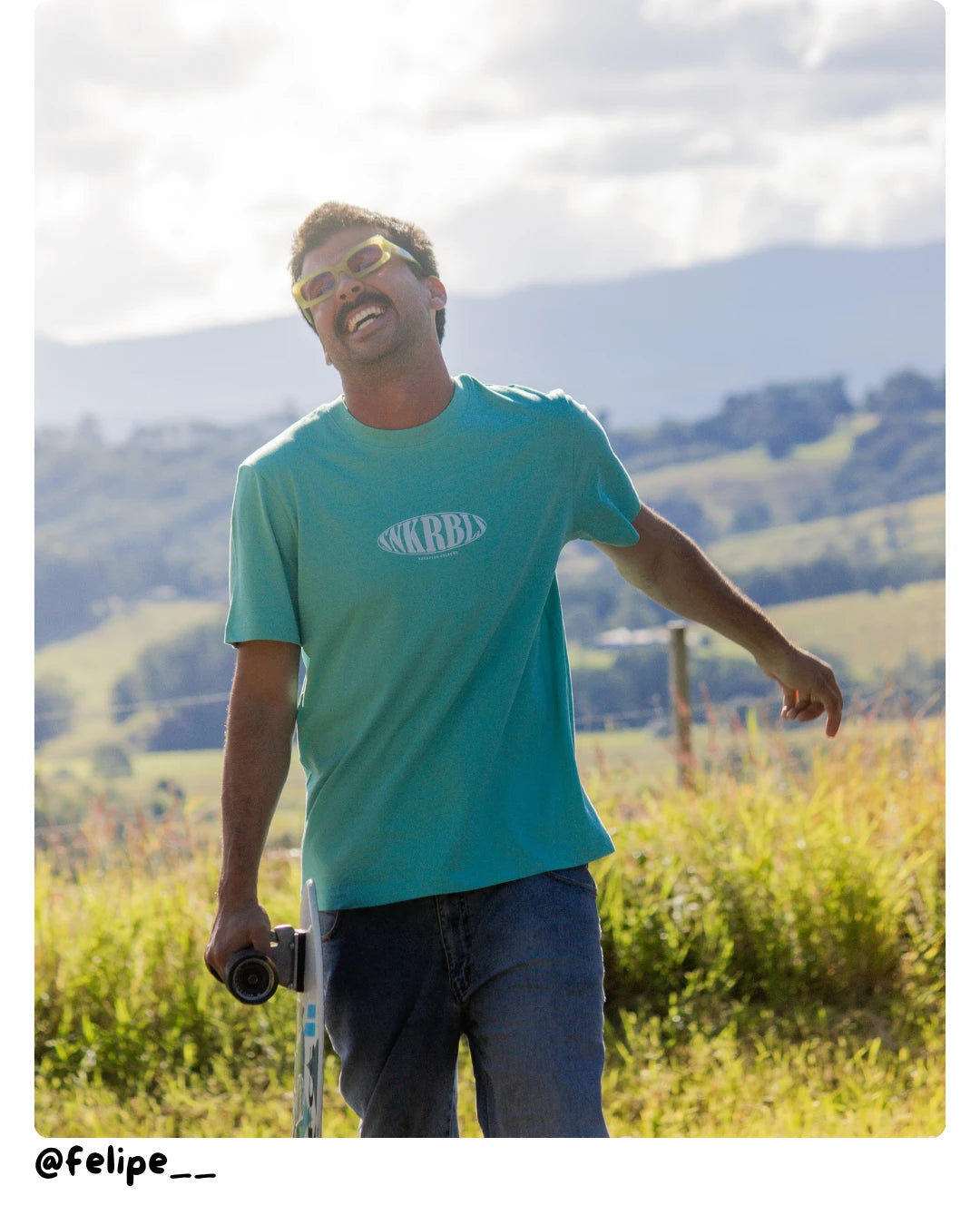 odel von The Sunkissed Rebel trägt ein türkisfarbenes T-Shirt und hält ein Skateboard in sommerlicher Landschaft.