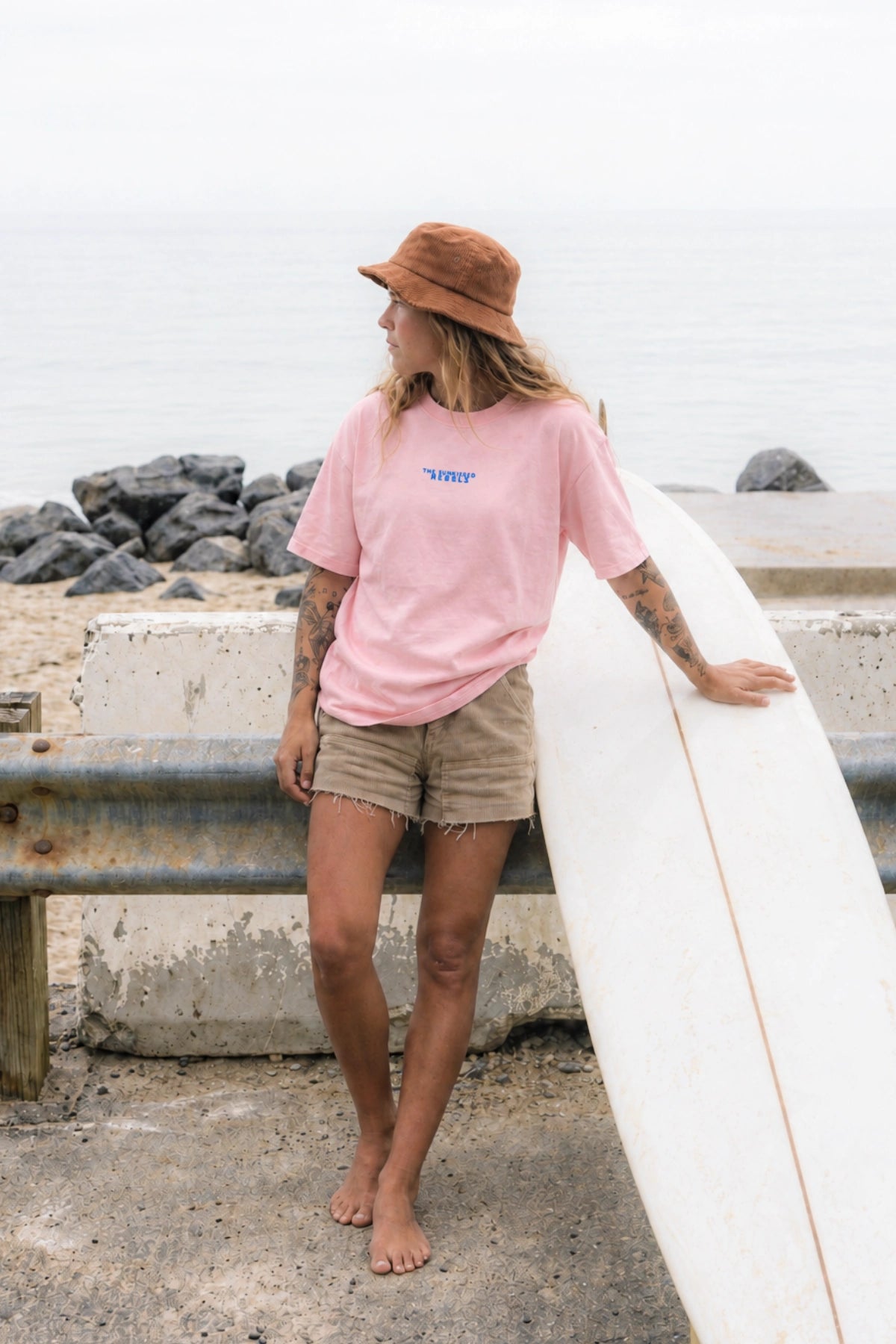 Frau barfuß mit Surfboard im rosa No Bullshit Whale Oversized T-Shirt mit blauem Brustprint, Bucket Hat und Shorts am Meer.
