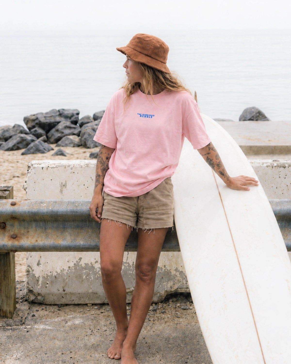 Frau barfuß mit Surfboard im rosa No Bullshit Whale Oversized T-Shirt mit blauem Brustprint, Bucket Hat und Shorts am Meer.