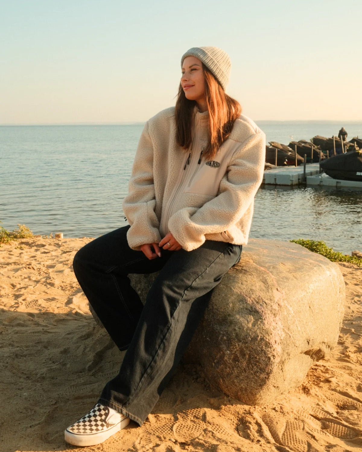 Frau am Strand mit beigefarbener Teddy Fleecejacke in Sand von The Sunkissed Rebels, Beanie und weiter Hose.