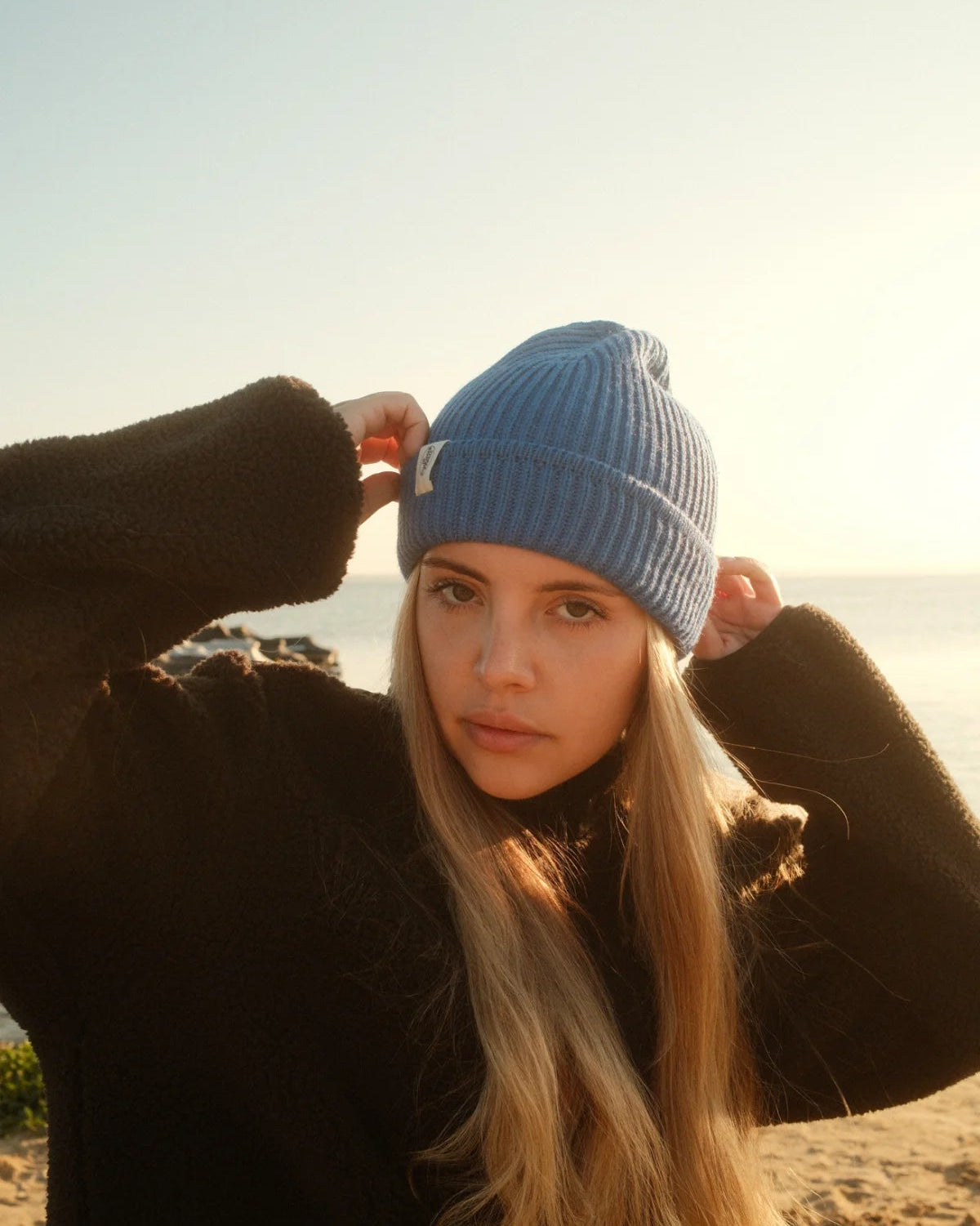 Entspannter Coastal Look mit der blauen Wollmütze Rebels Knit Beanie Ocean Blue von The Sunkissed Rebels und schwarzer Fleecejacke.