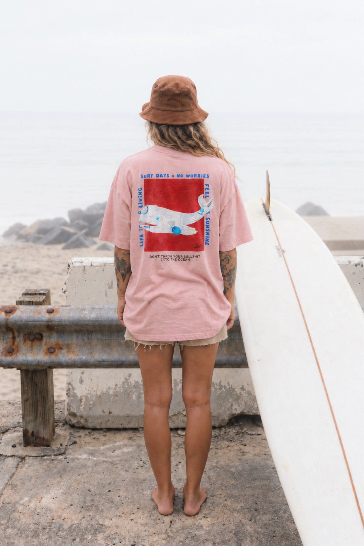 Surferin von hinten im rosa No Bullshit Whale Boxy Oversized T-Shirt von The Sunkissed Rebels mit Wal-Backprint neben Surfbrett am Strand.
