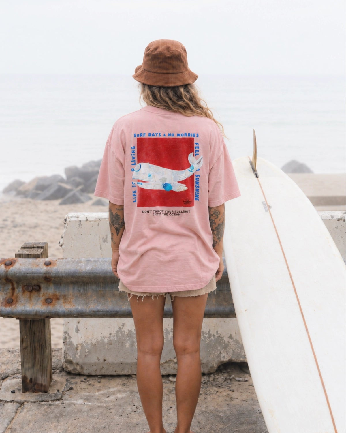 Surferin von hinten im rosa No Bullshit Whale Boxy Oversized T-Shirt von The Sunkissed Rebels mit Wal-Backprint neben Surfbrett am Strand.