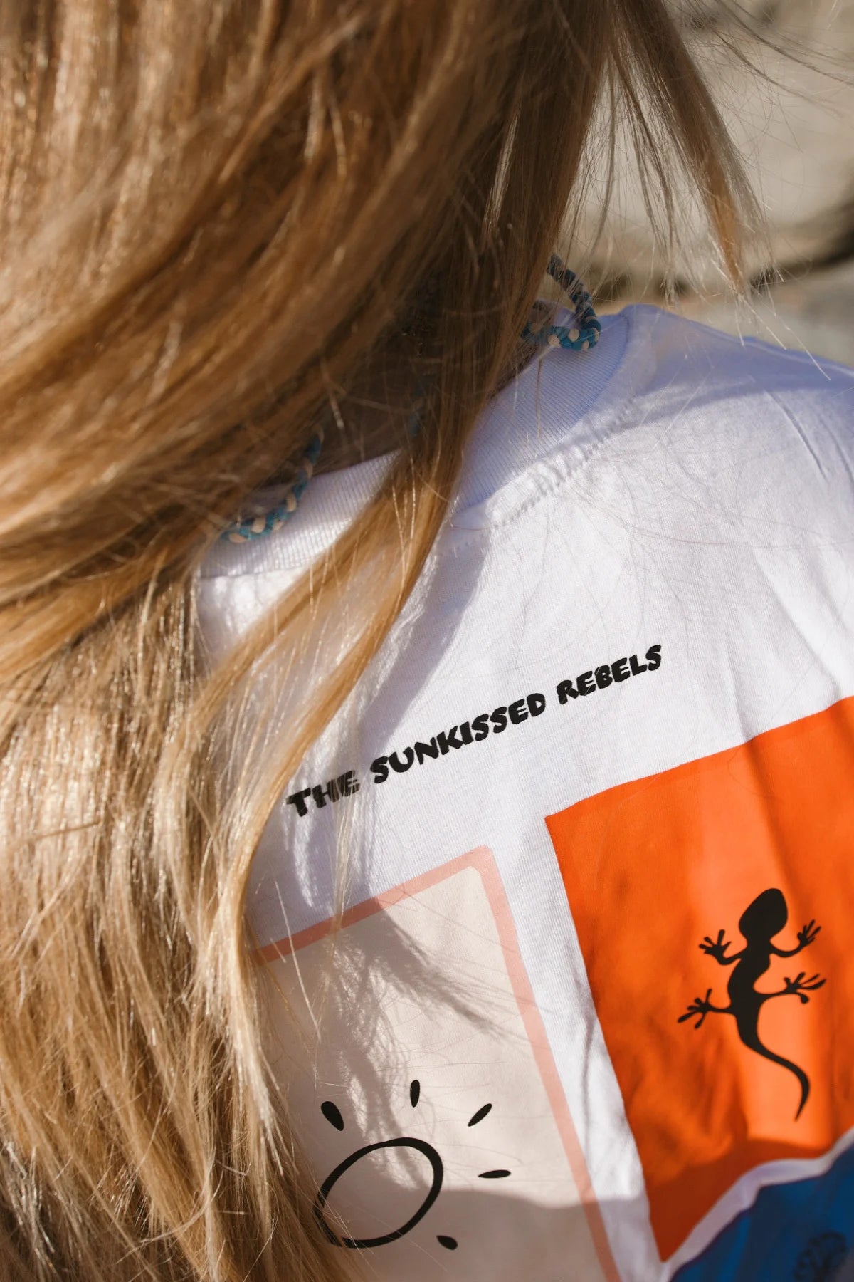 Detailansicht des Nackenbereichs und des oberen Rückenprints des weißen T-Shirts mit dem Markenschriftzug „The Sunkissed Rebels“.