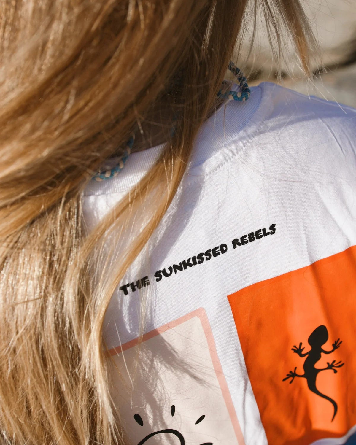 Detailansicht des Nackenbereichs und des oberen Rückenprints des weißen T-Shirts mit dem Markenschriftzug „The Sunkissed Rebels“.
