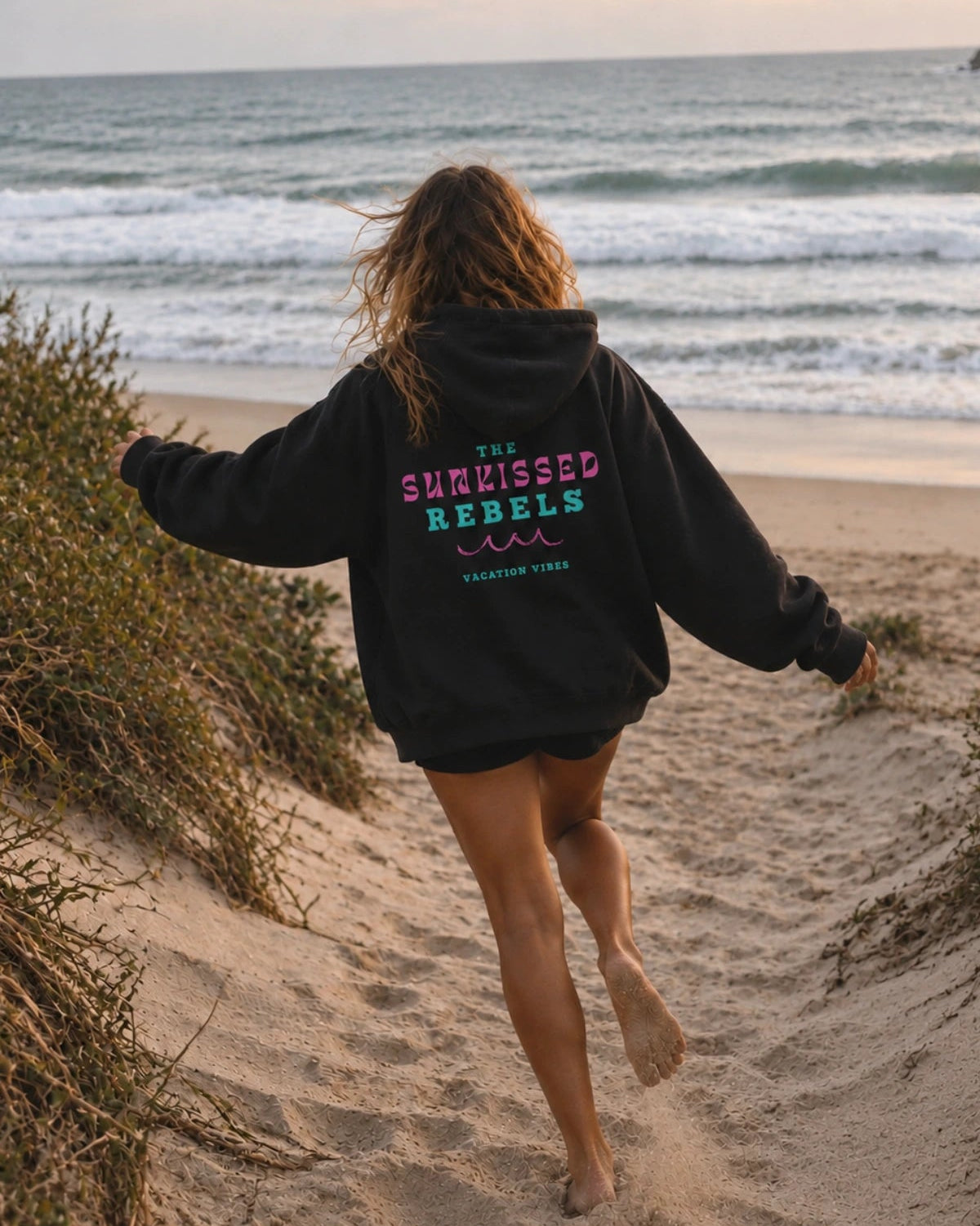 Frau barfuß im schwarzen Vacation Vibes Hoodie am Strand Richtung Meer laufend – The Sunkissed Rebels