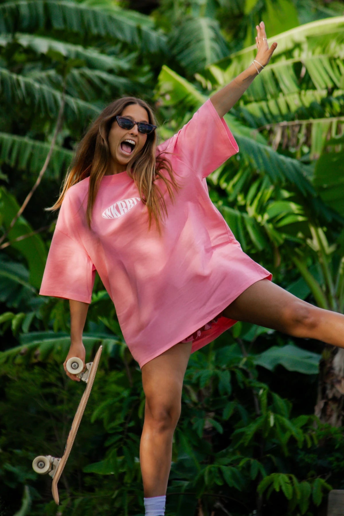 Frau mit Skateboard in dynamischer Pose im SNKRBLS Pink Joy Oversized T-Shirt vor tropischem Hintergrund.