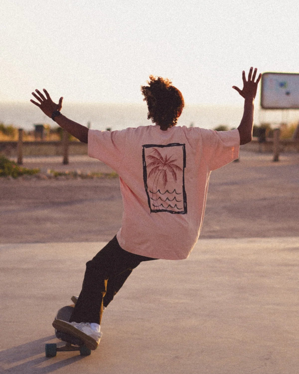 Skater in Korallfarbenem Oversized T-Shirt von The Sunkissed Rebels mit Palmendruck bei Sonnenuntergang