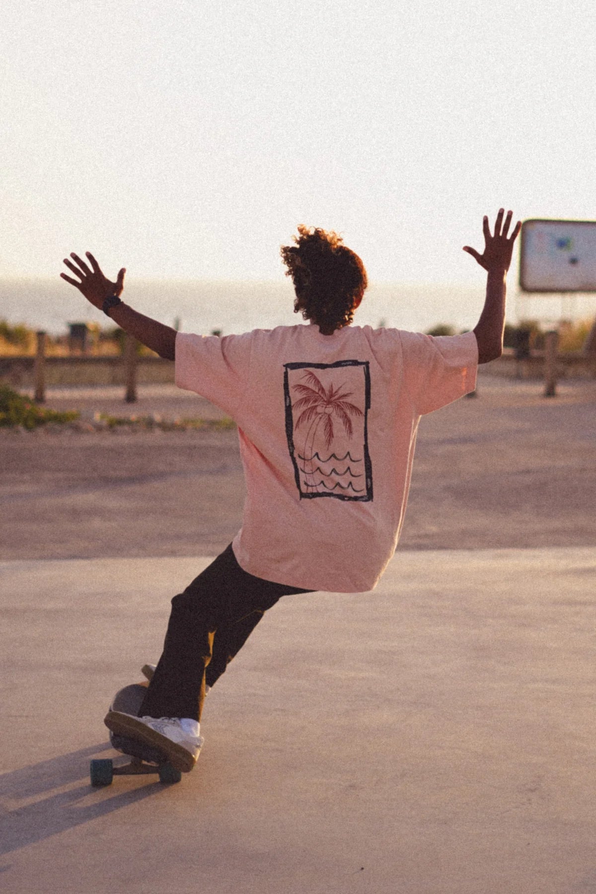 Skater in Korallfarbenem Oversized T-Shirt von The Sunkissed Rebels mit Palmendruck bei Sonnenuntergang