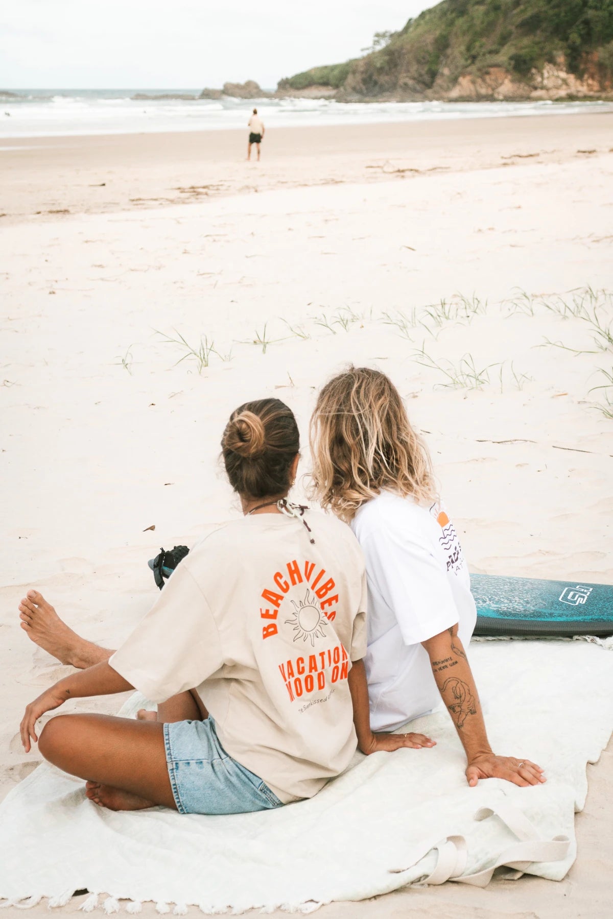 Pärchen mit The Sunkissed Rebels T-Shirts am Strand mit Surfbrett – Boho & Surfstyle