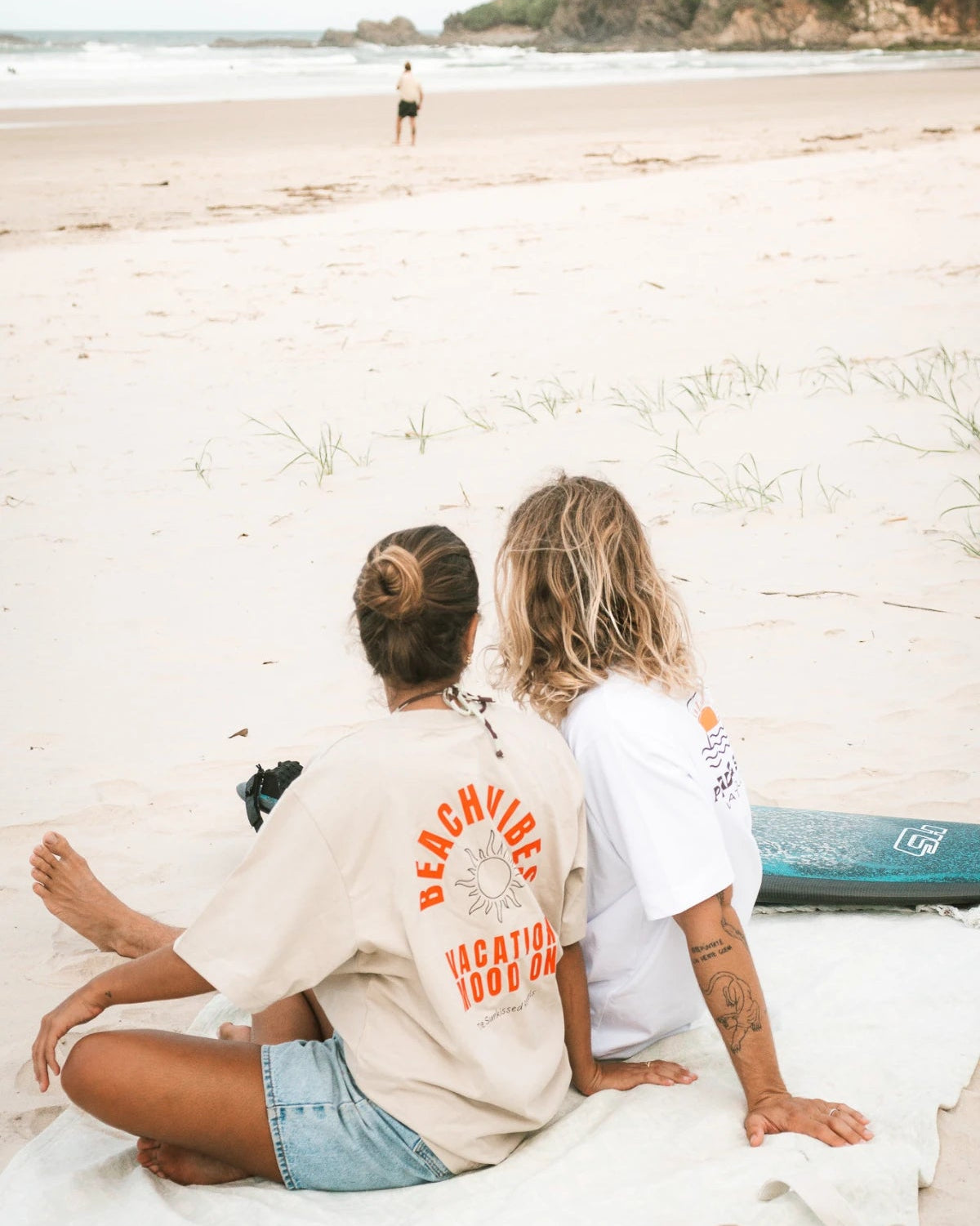 Pärchen mit The Sunkissed Rebels T-Shirts am Strand mit Surfbrett – Boho & Surfstyle