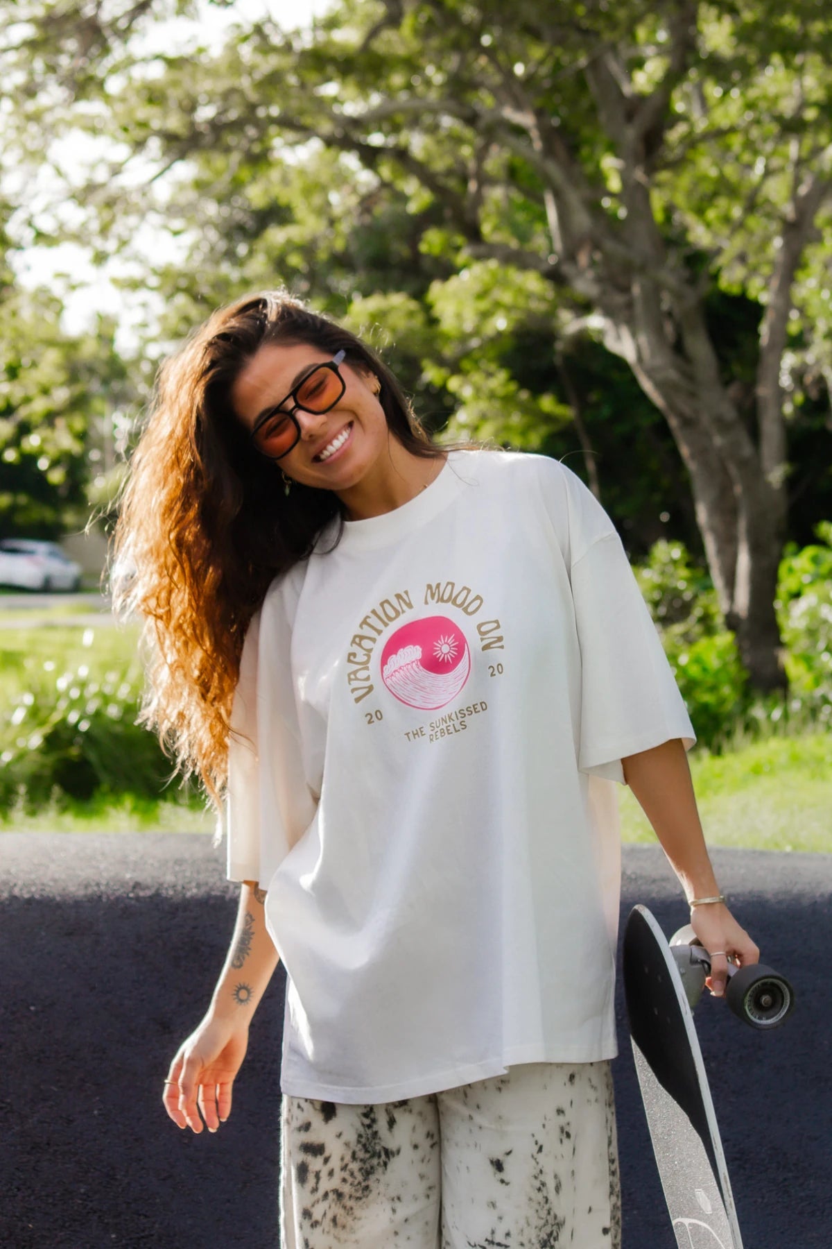 Frau im Cream Oversized T-Shirt Vacation Mood On von The Sunkissed Rebels mit Skateboard im Sonnenlicht