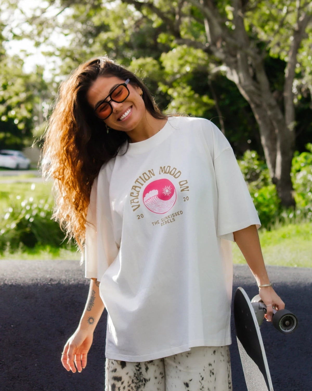 Frau im Cream Oversized T-Shirt Vacation Mood On von The Sunkissed Rebels mit Skateboard im Sonnenlicht