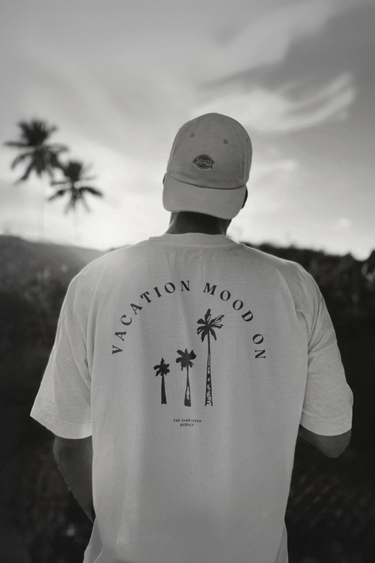Rückenansicht eines Mannes im weißen 'Vacation Mood On' T-Shirt vor Sonnenuntergang und Palmen