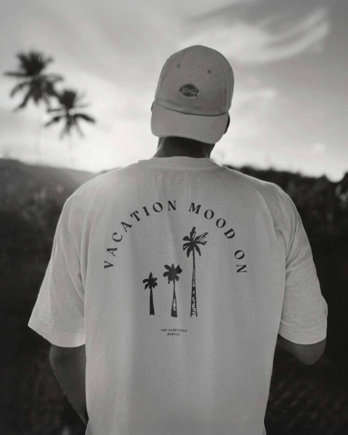 Rückenansicht eines Mannes im weißen 'Vacation Mood On' T-Shirt vor Sonnenuntergang und Palmen