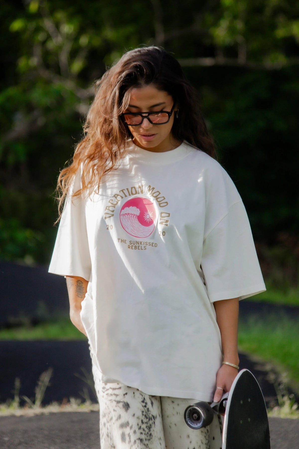 Frau im The Sunkissed Rebels Cream T-Shirt Vacation Mood On steht nachdenklich mit Skateboard