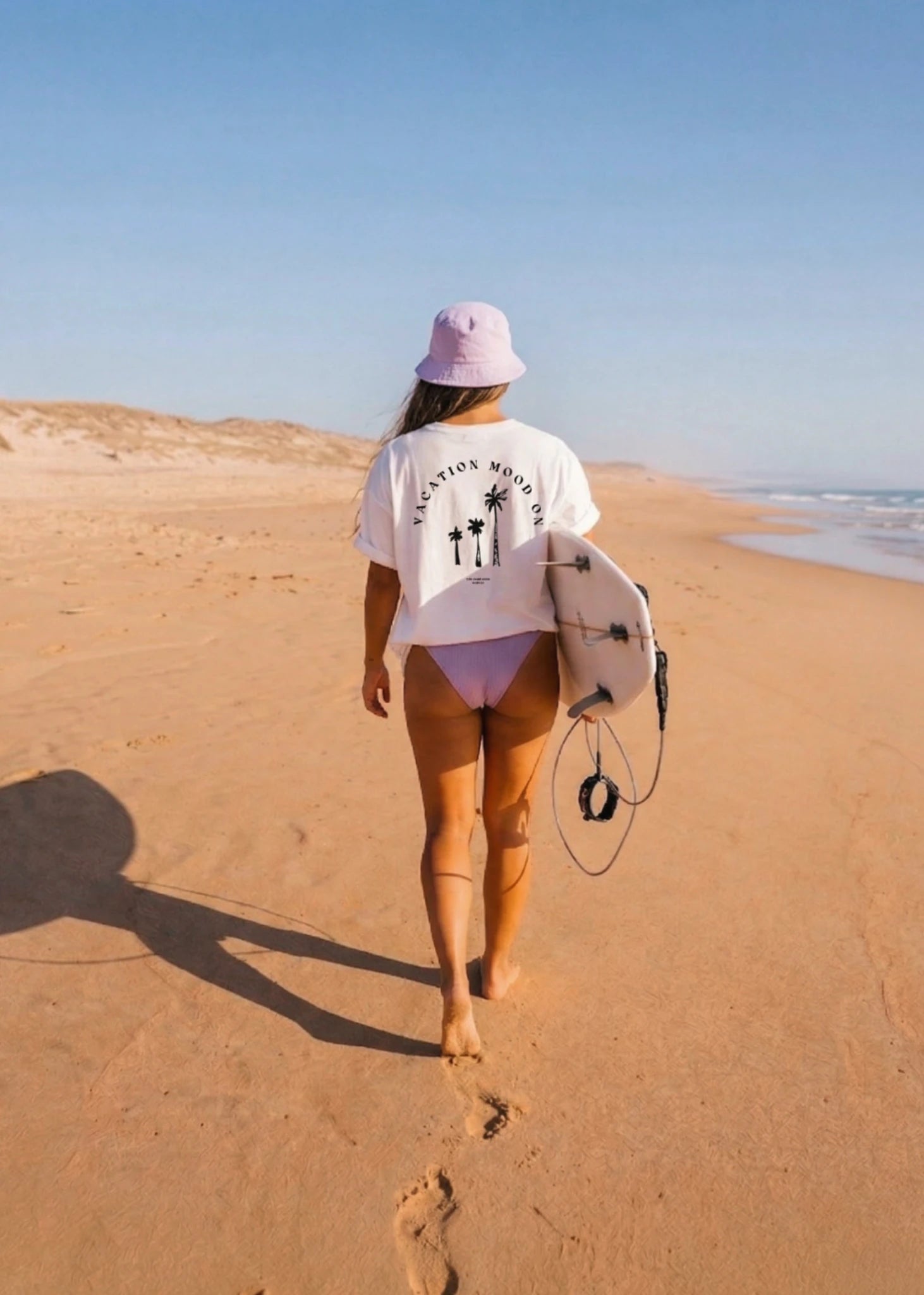 Rückansicht einer Frau am Strand mit Surfboard trägt weißes Vacation Mood On T-Shirt mit Rückenprint – entspannter Sommer Look