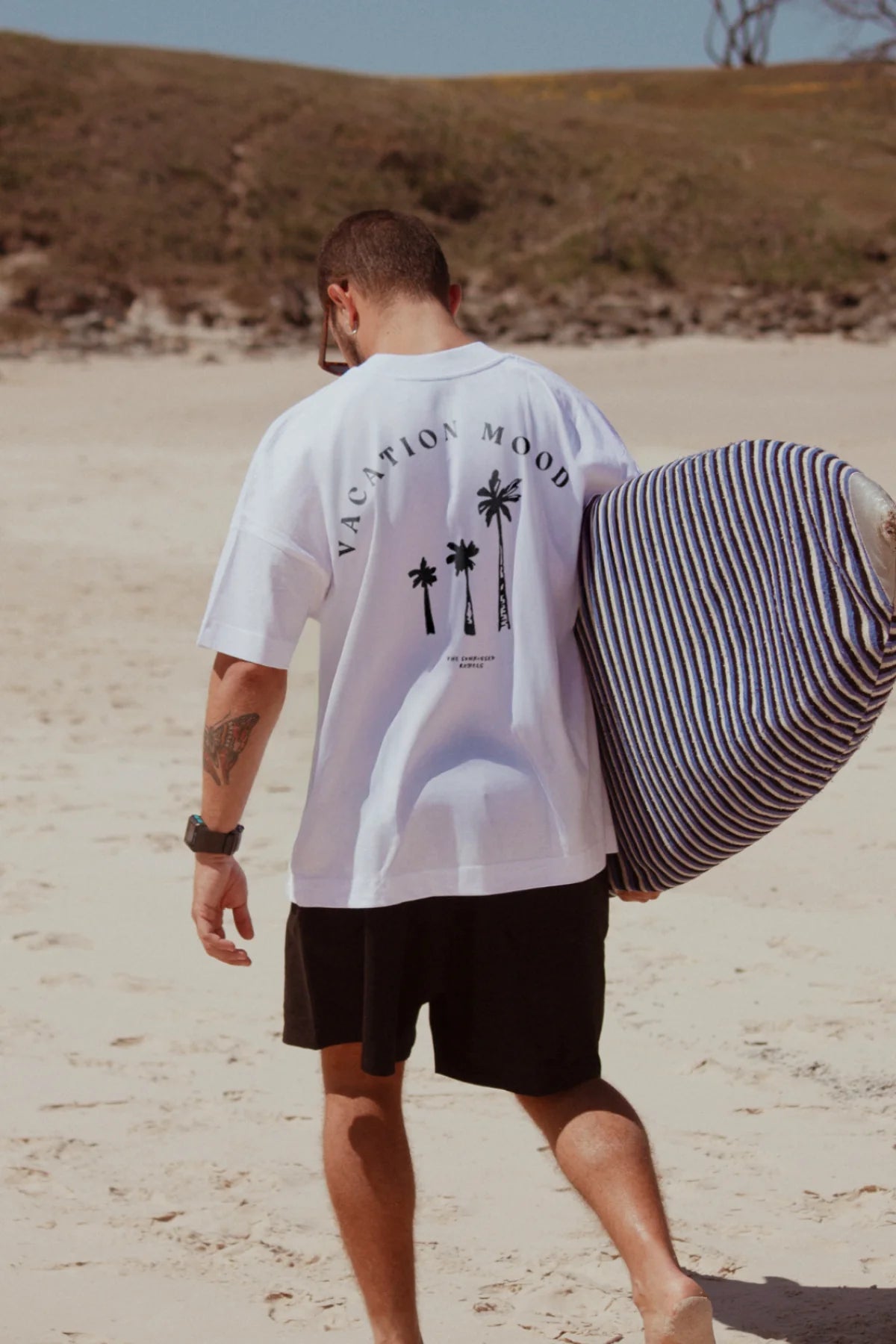Rückansicht eines Mannes im weißen Oversized T-Shirt mit 'Vacation Mood On'-Print am Strand
