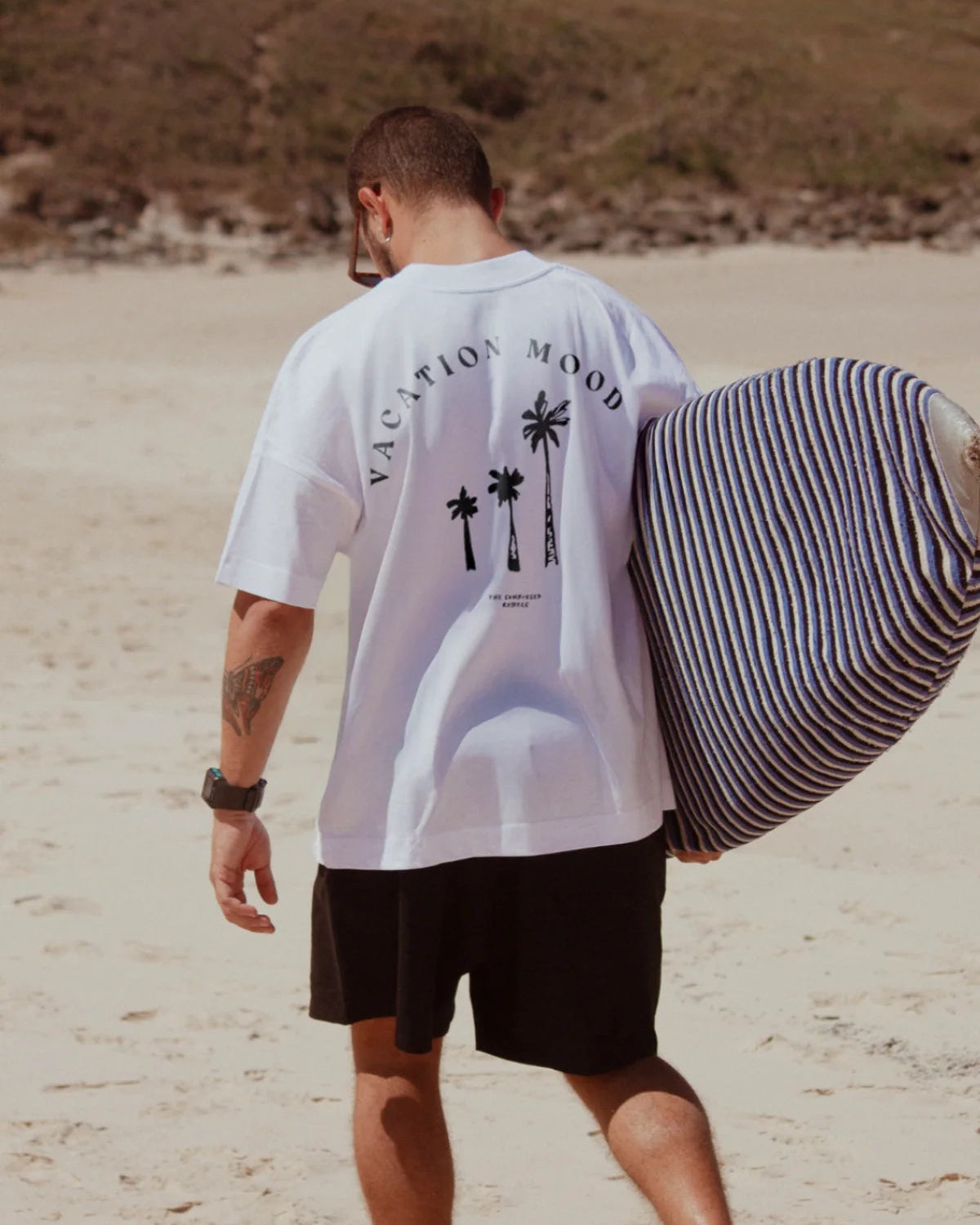 Rückansicht eines Mannes im weißen Oversized T-Shirt mit 'Vacation Mood On'-Print am Strand