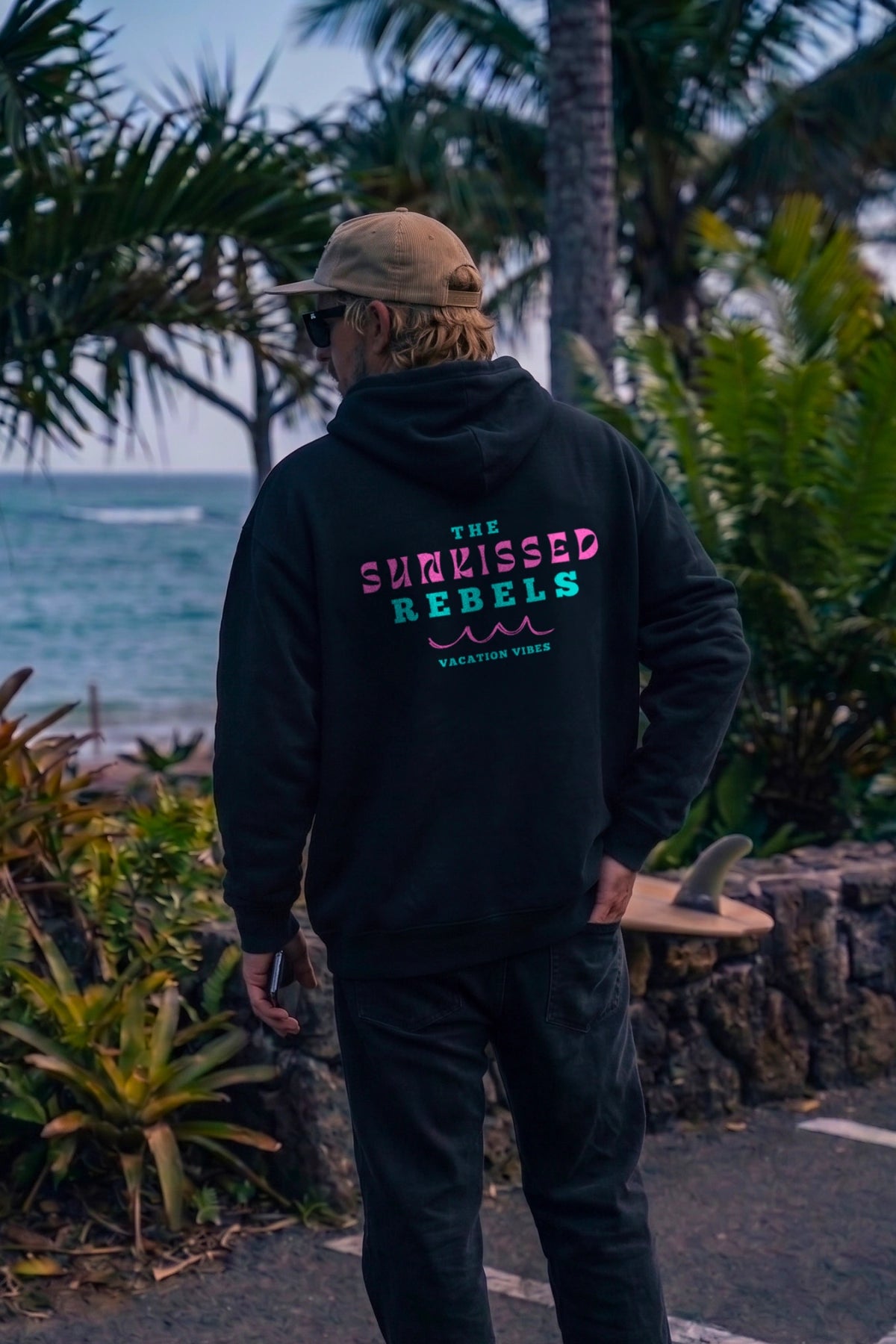 Männliches Model präsentiert den schwarzen Vacation Vibes Oversized Hoodie von The Sunkissed Rebels mit dezentem Frontlogo vor tropischer Strandkulisse.