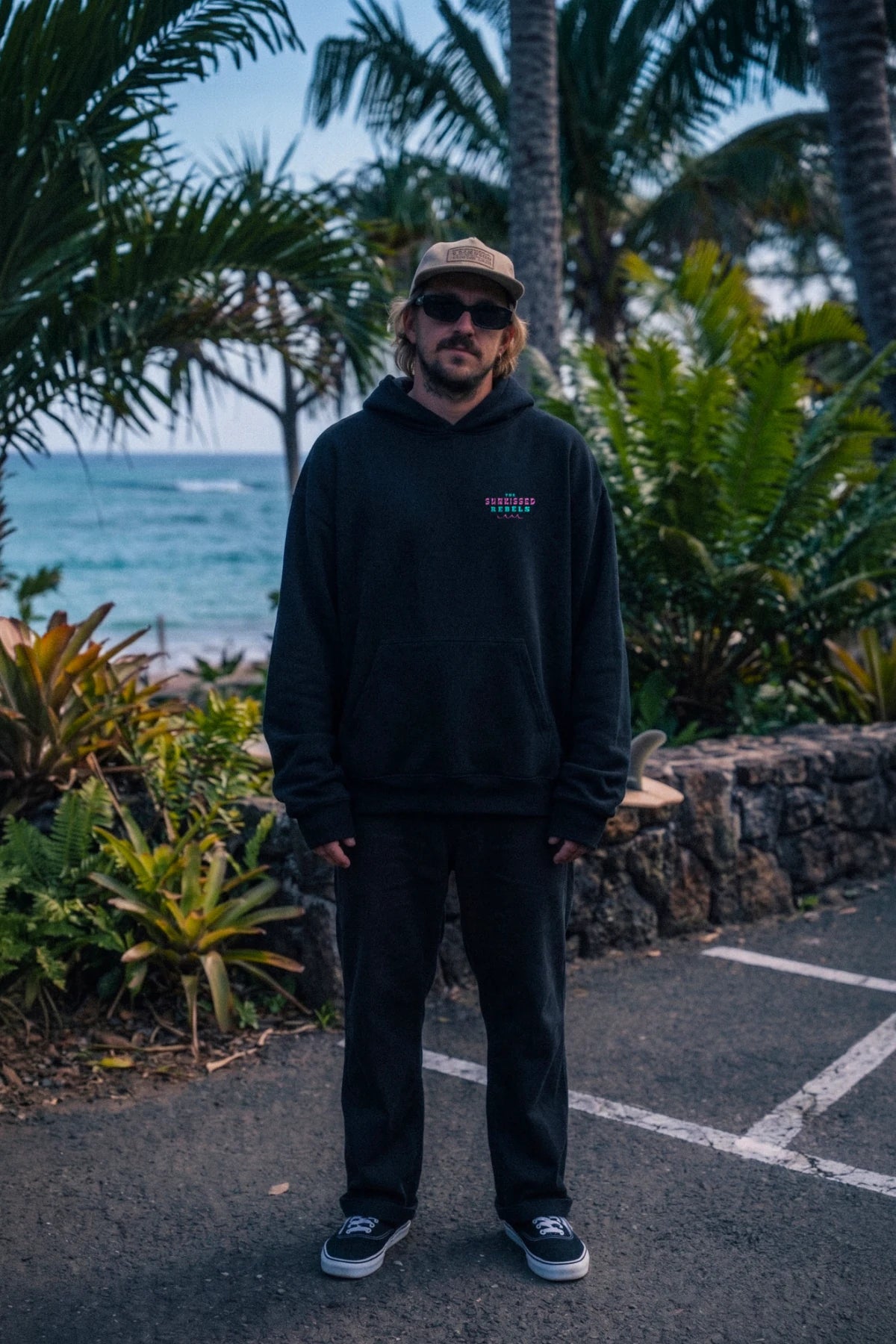 Männliches Model trägt den schwarzen Vacation Vibes Oversized Hoodie von The Sunkissed Rebels mit großem Backprint in tropischer Küstenkulisse.