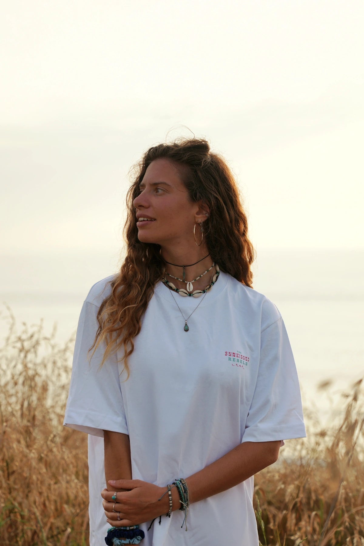 Frau mit Muschelketten am Ozean trägt weißes Vacation Vibes T-Shirt – dezenter Print auf der Brust.