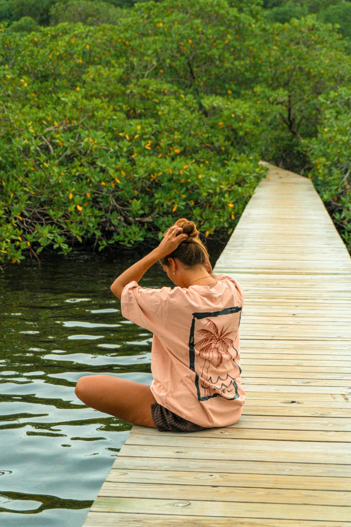 Rückseite des Waves & Palm Trees Oversized T-Shirts in Coral mit großem Palmen-Backprint, getragen von einer Frau auf einem Holzsteg am Wasser.