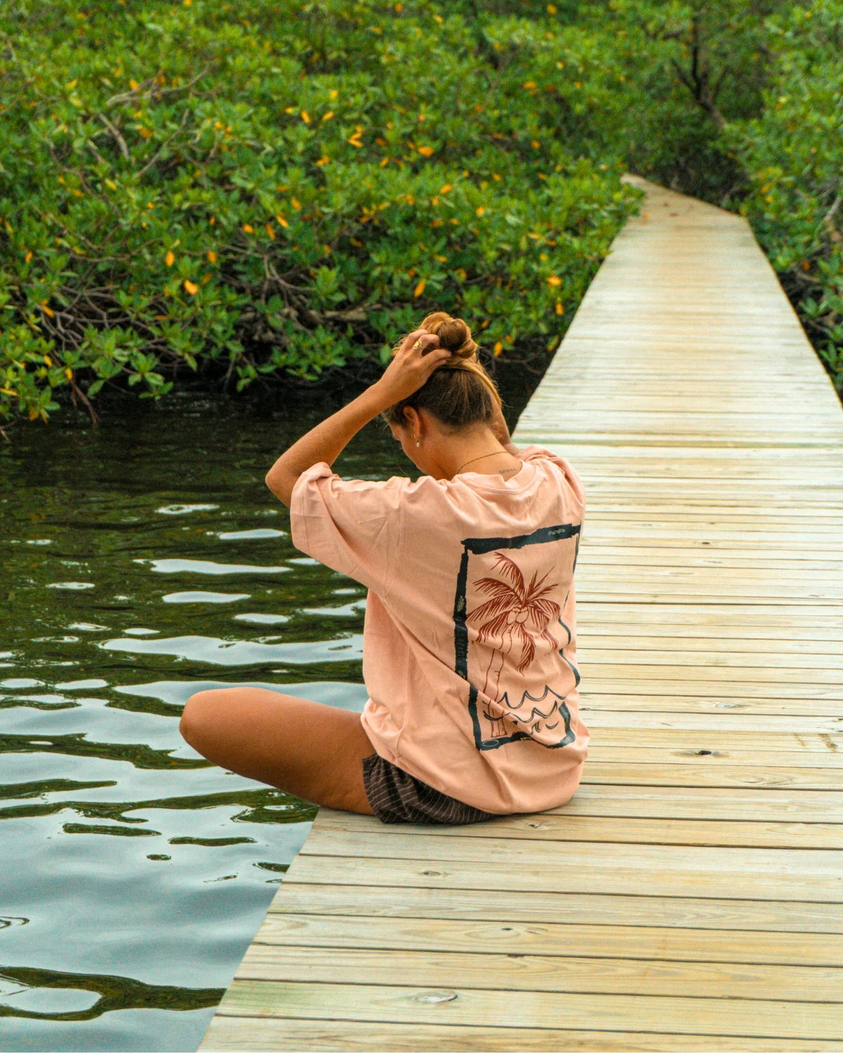 Rückseite des Waves & Palm Trees Oversized T-Shirts in Coral mit großem Palmen-Backprint, getragen von einer Frau auf einem Holzsteg am Wasser.